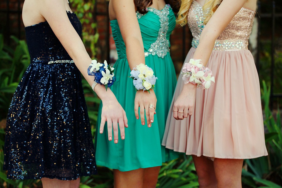 Three girls wearing prom dresses holding out their arms with corsages on them.