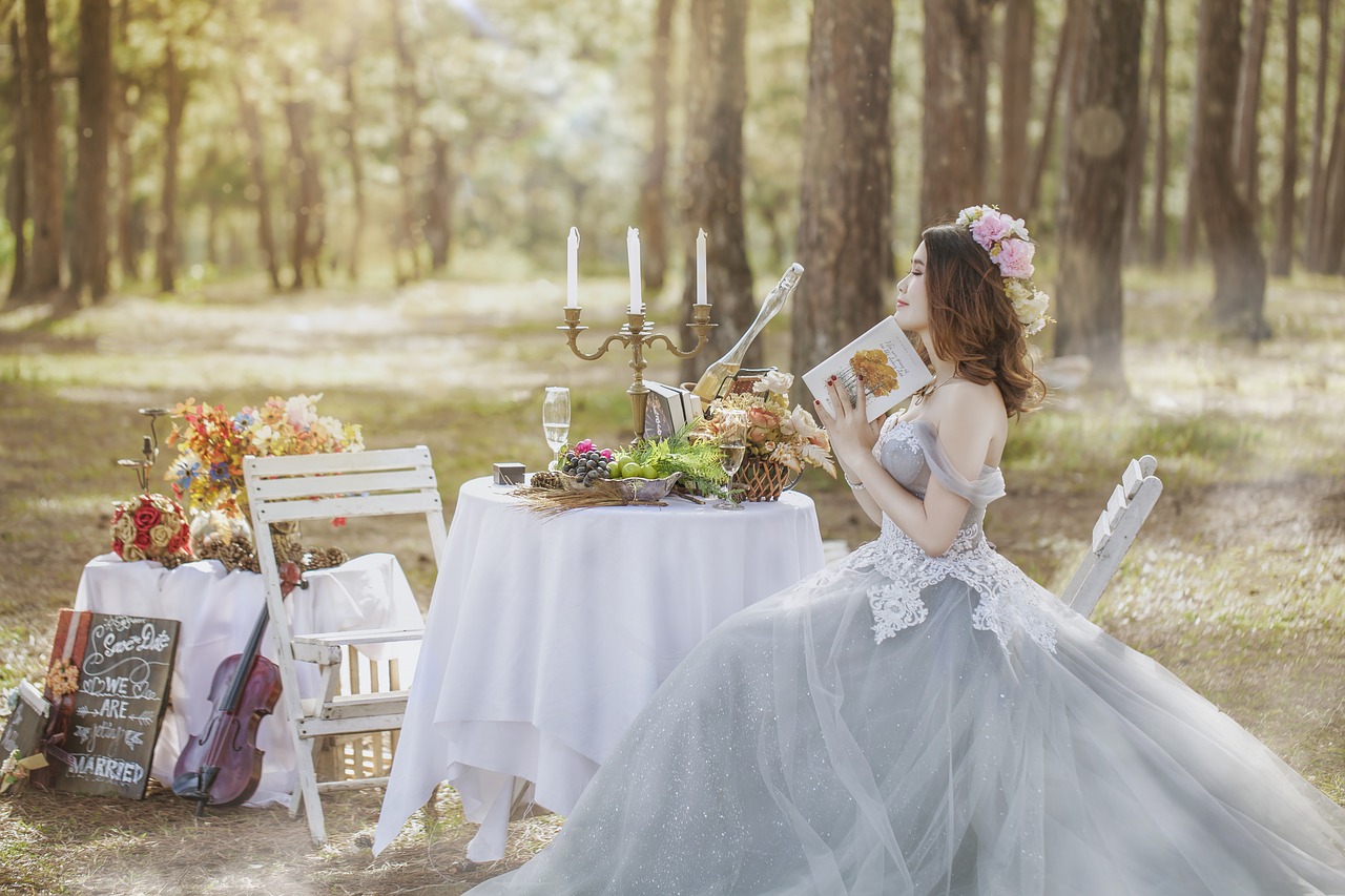 Smiling bride holding a book sitting in the forest at a food and drink laden table.
