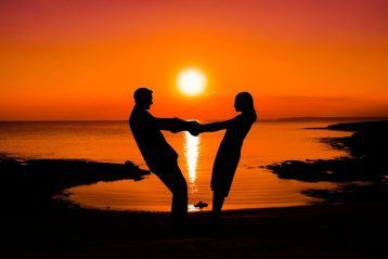 A couple standing on the beach holding hands while the sun rises.