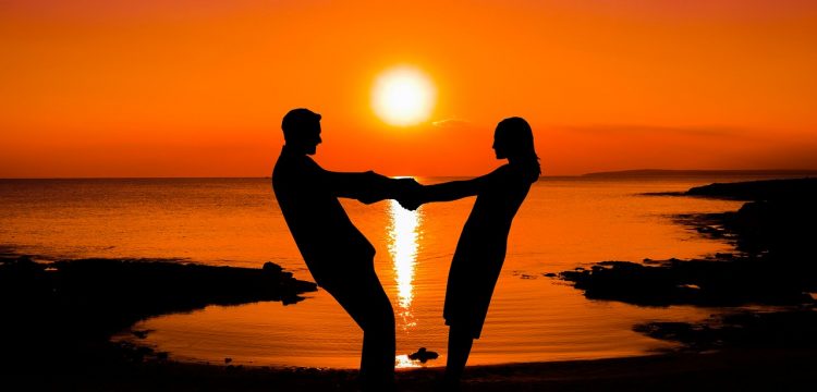 A couple standing on the beach holding hands while the sun rises.