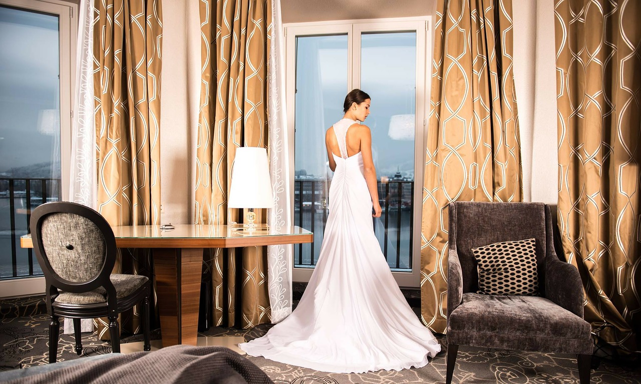 Woman wearing wedding dress in an elegant room, looking out a window.