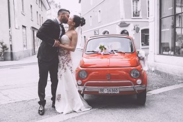 Bride and groom kissing by an orange sports car.